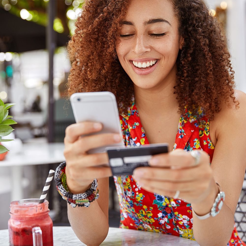 1beautiful-dark-skinned-young-female-with-cheerful-expression-holds-smart-phone-credit-card-banks-online-makes-shopping-while-sits-against-cafe-interior_web